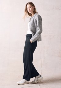 Šedá strukturovaná bunda, bílý tričko, černé volné kalhoty a bílé tenisky se šedými akcenty. Model stojí, ruce v kapsách.