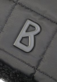Schwarze Stofftasche mit gesteppter Textur, die ein schwarzes Gummi-Logo in Form eines 'B' trägt. Der Boden hat einen samtigen schwarzen Akzent für den Kontrast.
