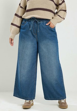 Person trägt weite blaue Jeans mit Kordelzug in der Taille, beigen gestreiften Pullover und beige Sneaker mit weißen Schnürsenkeln.