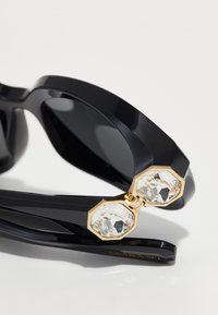 Ochelari de soare negri cu un cadru gros, având accente aurii și două cristale mari și clare la balamale. Temelia include marca "SWAROVSKI".