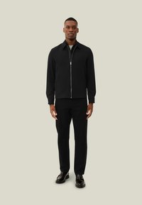Giacca nera con zip e maniche lunghe, indossata sopra pantaloni neri e scarpe nere. Texture liscia, design minimalista, senza motivi visibili.