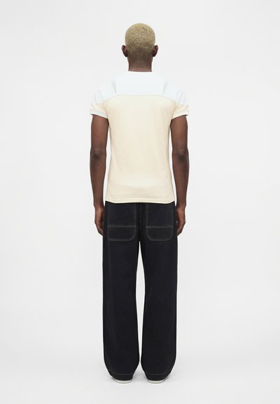 Άτομο με κοντά ξανθά μαλλιά στέκεται με την πλάτη προς το μέρος μας, φοράει μπλουζάκι με κοντά μανίκια σε μπεζ και άσπρο χρώμα και φαρδιά μαύρη παντελόνια με τσέπες.