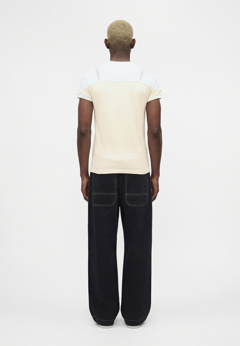 Personne aux cheveux blonds courts, de dos, portant une chemise beige et blanche à manches courtes et un pantalon noir ample avec des poches plaquées.