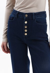 Tmavě modré džíny z denimu s pěti předními knoflíky, vysokým pasem a dvěma bočními kapsami. Látka má hladký, mírně strukturovaný povrch.