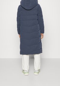 Marineblå puffjakke med hætte, med horisontale quiltede sektioner, kombineret med hvide bukser og grønne sneakers.