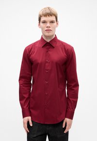 Giovane uomo con i capelli corti, che indossa una camicia bordeaux a maniche lunghe con bottoni e pantaloni scuri, su uno sfondo bianco uniforme.