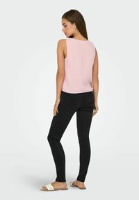 Lichtroze mouwloos topje van een gestructureerde stof, gecombineerd met zwarte skinny jeans en witte platte sandalen. Model gezien vanuit de rug.