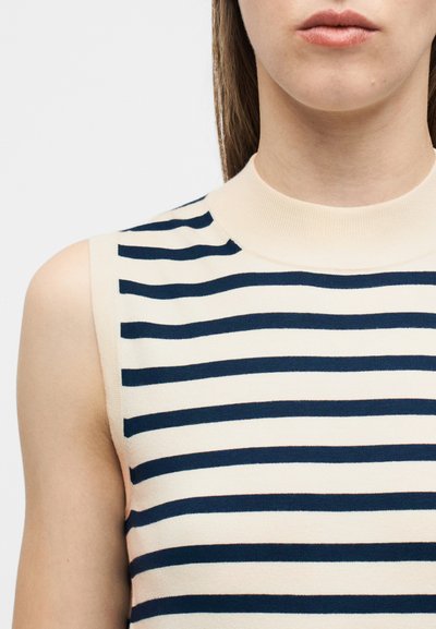 Κοντινό πλάνο ενός ατόμου που φοράει μπεζ αμάνικο τοπ με οριζόντιες ναυτικές μπλε ρίγες και ψηλό στρογγυλό ντεκολτέ.