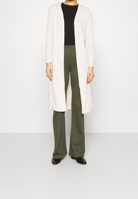 Cardigan largo y acanalado en crema, diseño de frente abierto, combinado con una blusa negra y pantalones en verde oliva, todo sobre un fondo neutro.