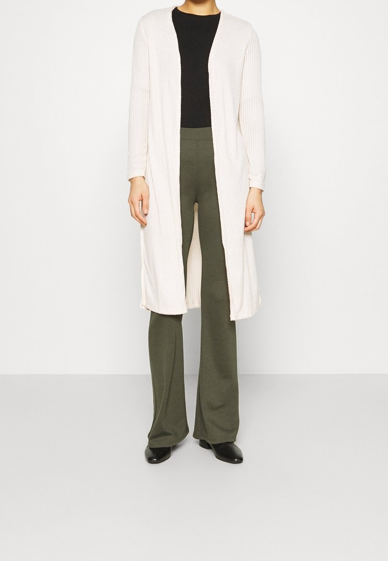 Cardigan largo y acanalado en crema, diseño de frente abierto, combinado con una blusa negra y pantalones en verde oliva, todo sobre un fondo neutro.