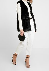 Svart faux fur-väst med öppen front, bärs över en ljusfärgad polotröja. Matchas med vita cropped-jeans och svarta sandaler med remmar. Rund svart handväska.