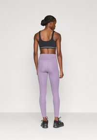 Μαύρο αθλητικό σουτιέν με ριγωτή ταινία, συνδυασμένο με κολάν υψηλής μέσης σε ανοιχτό μοβ χρώμα. Το μοντέλο φοράει μαύρα αθλητικά παπούτσια.