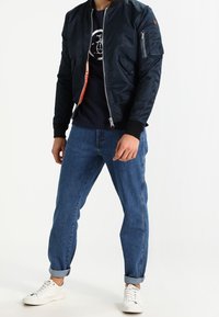 Homme portant une veste bomber noire, un t-shirt graphique sombre, un jean bleu retroussé et des baskets blanches sur fond blanc.
