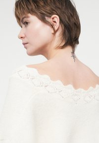 Cremfarbener Strickpullover mit einem gewellten Spitzenkragen; weiche Textur und lange Ärmel. Auf dem Rücken ist ein florales Tattoo am Hals zu sehen.