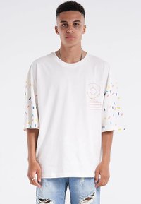 Weißes übergroßes T-Shirt mit bunten Streuselmuster auf den kurzen Ärmeln. Auf der linken Brustseite vorne befindet sich ein kleines rosa Design.