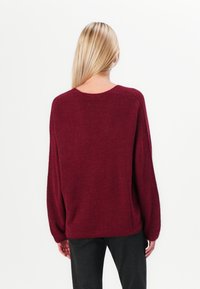 Maglione a maglia bordeaux con una vestibilità ampia, scollo rotondo e maniche lunghe. Texture morbida e dettaglio a coste sull'orlo. Indossato sopra pantaloni neri a righe.