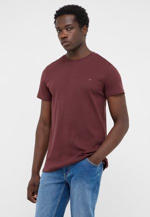 Junger Mann mit dunklen lockigen Haaren, der ein bordeauxfarbenes kurzärmeliges T-Shirt und blaue Jeans trägt, steht mit einer Hand in der Tasche.
