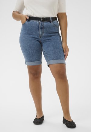 Osoba ubrana w niebieskie szorty z denimu z podwiniętymi nogawkami i czarnym paskiem, biały krótki top z krótkim rękawem oraz czarne baleriny, stojąca z jedną ręką w kieszeni.