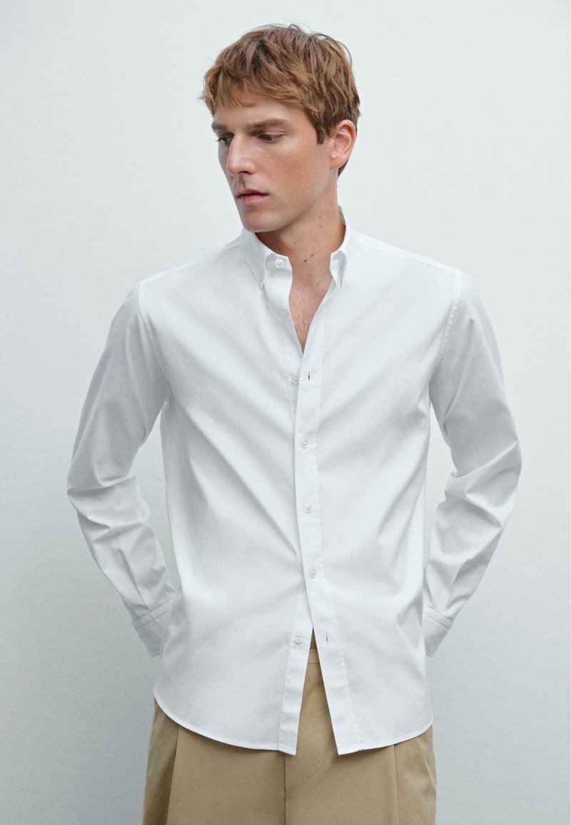 Jeune homme aux cheveux châtain clair portant une chemise blanche à boutons et un pantalon beige, regardant légèrement vers sa gauche sur un fond uni.