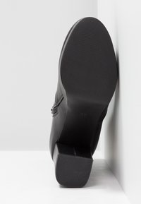 Bottine noire à la cheville avec la semelle reposant contre un mur blanc, montrant un talon texturé et une semelle lisse avec la taille 37 marquée sur la semelle.