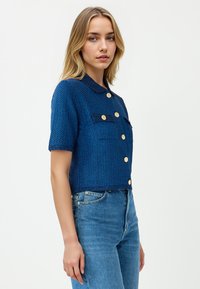 Junge Frau mit blonden Haaren, die eine strukturiertes blaues Kurzarm-Button-Up-Cardigan und hochgeschnittene blaue Jeans trägt und seitlich steht.