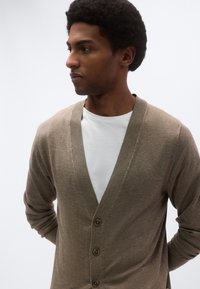 Καφέ ζακέτα με βαθύ V λαιμόκοψη, ριγωτές άκρες και πέντε κουμπιά. Φορεμένη πάνω από λευκό T-shirt, με ομαλή, απαλή υφή.