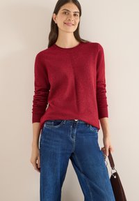 Maglione rosso con polsini e collo a coste, caratterizzato da un design con cucitura centrale. Abbinato a jeans blu a gamba larga e una borsa a spalla marrone.