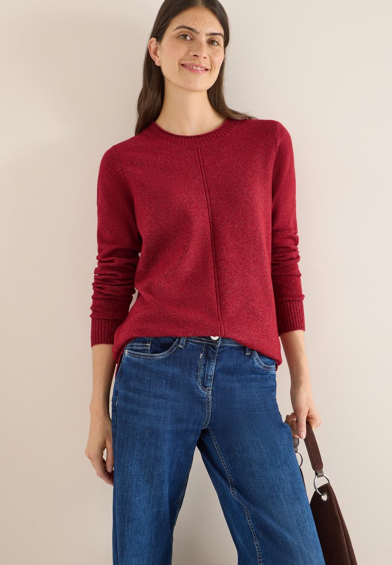 Maglione rosso con polsini e collo a coste, caratterizzato da un design con cucitura centrale. Abbinato a jeans blu a gamba larga e una borsa a spalla marrone.
