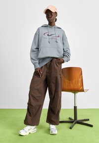 Grauer Kapuzenpulli mit rotem Nike-Logo, lockere braune Cargo-Hosen und grau-weiße Sneaker mit sichtbarer Luftpolsterung. Weiche Texturen und lässige Passform.