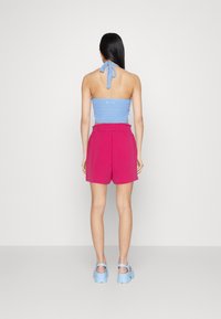 Halterowy top na szydełku w jasnoniebieskim kolorze, zestawiony z różowymi szortami z wysokim stanem. Modelka ma na sobie masywne niebieskie sandały. Gładkie białe tło.