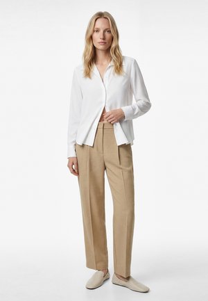 Femme blonde portant une chemise blanche boutonnée, un pantalon beige tailleur et des chaussures beige à enfiler, debout devant un fond blanc uni.