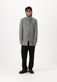 Cappotto grigio in misto lana con colletto alto, chiusura a bottoni e tasche laterali. Abbinato a pantaloni neri e scarpe nere, indossato con occhiali da sole.
