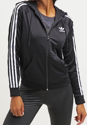 Czarna bluza Adidas na zamek z białymi paskami na rękawach, przednimi kieszeniami oraz logo na piersi, noszona na ciemnej kwiecistej koszuli.