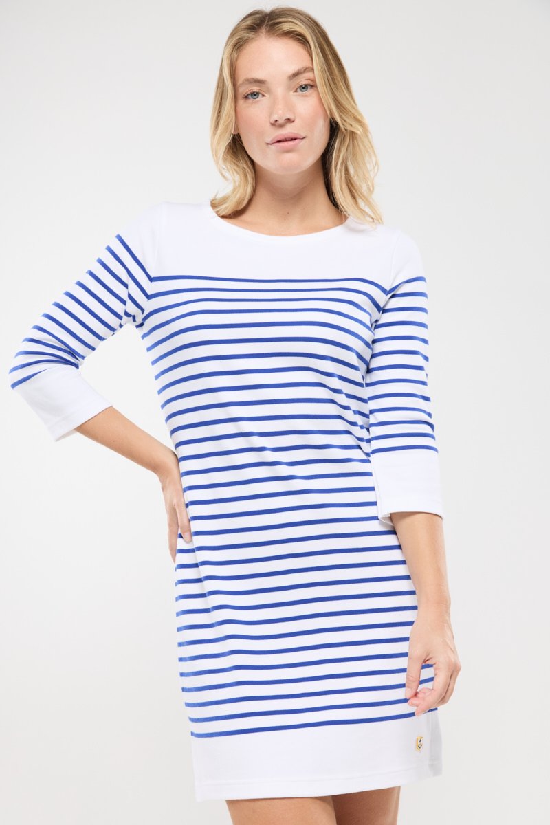 Femme blonde portant une robe blanche à rayures horizontales bleues et des manches trois-quarts, se tenant avec une main sur la hanche.