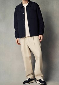 Námořnická texturovaná bunda se dvěma náprsními kapsami, bílé tričko, krémové plisované kalhoty a černé tenisky s bílými tkaničkami a krémovobílou podešví.