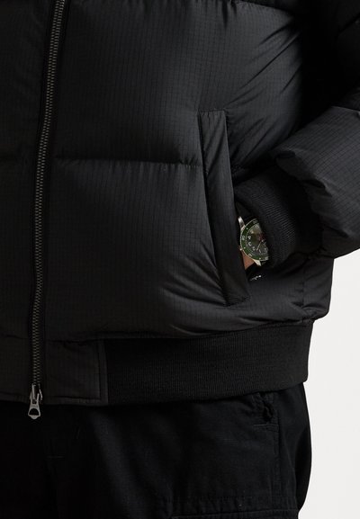 Čierna prešívaná bunda s prešívanou textúrou, predným zipsom, rebrovanými manžetami a bočnou vreckom. Na zápästí je čiastočne viditeľné zelené hodinky.