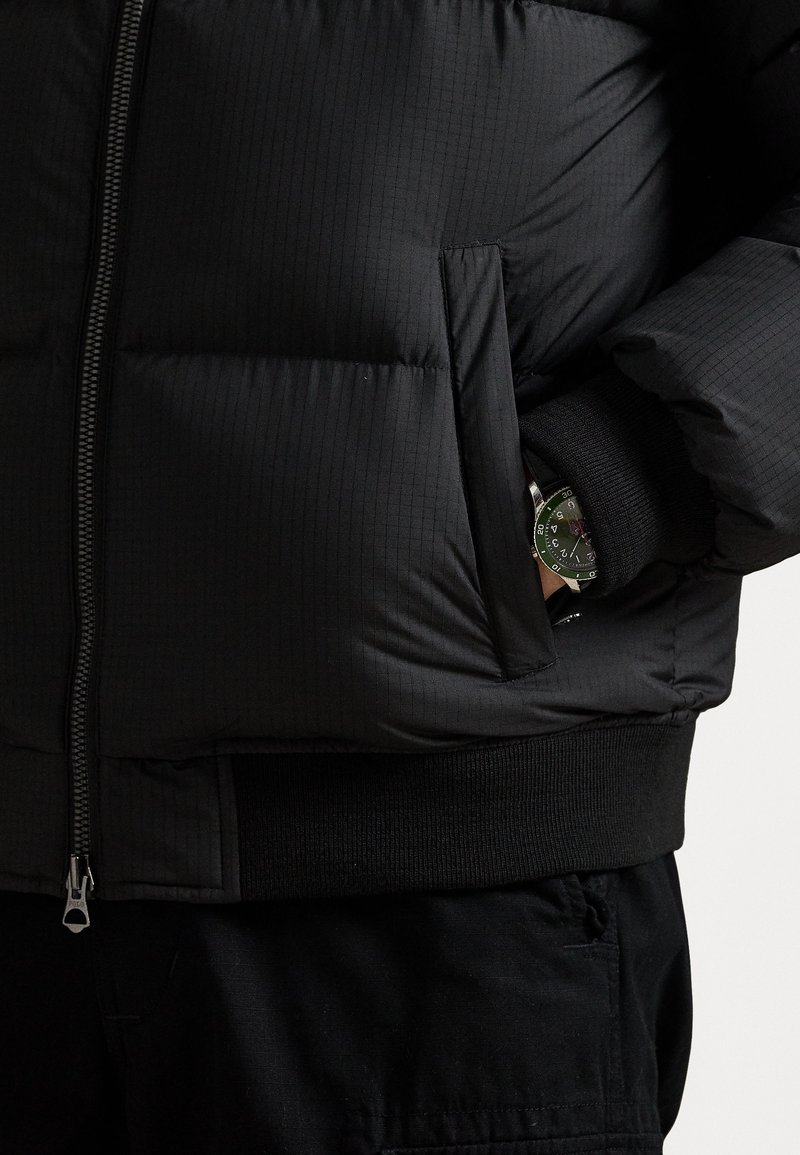 Zwarte pufferjack met gequiltde textuur, voorrits, ribgebreide manchetten en een zijzak. Een groene horloge is gedeeltelijk zichtbaar op de pols.