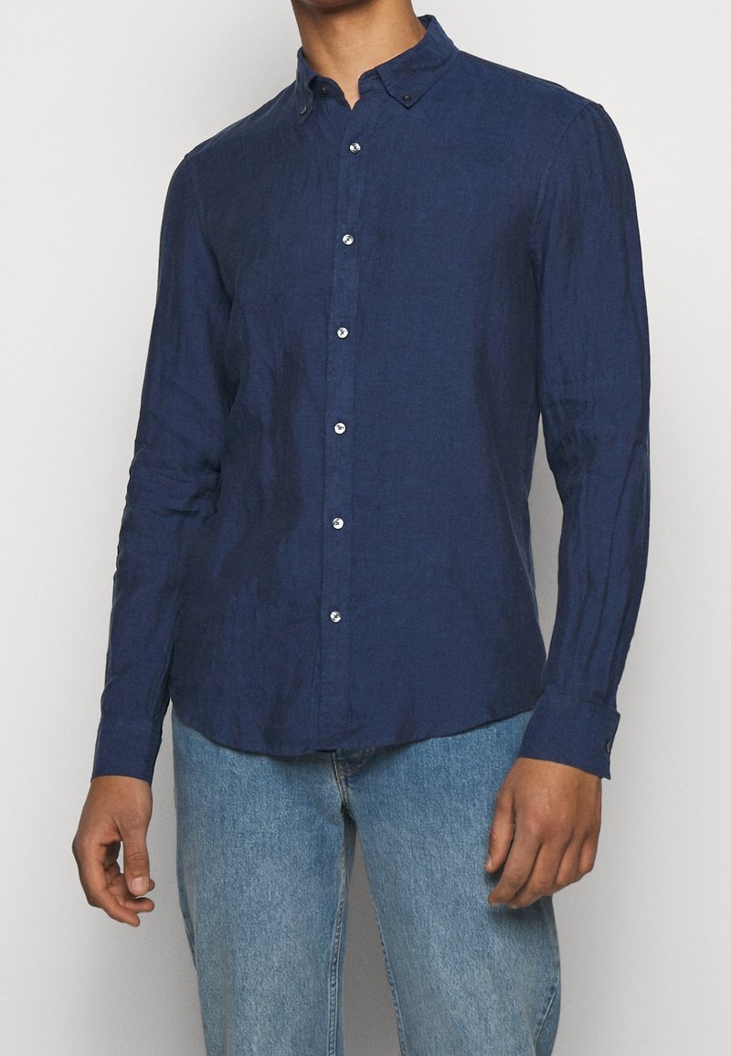 Homme portant une chemise ajustée à manches longues bleu marine à boutons, avec un jean bleu clair, sur un fond uni.
