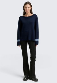 Maglione a maglia blu navy con maniche lunghe e collo ampio; presenta polsini a motivo blu. Abbinato a pantaloni neri a zampa d'elefante e scarpe scure.