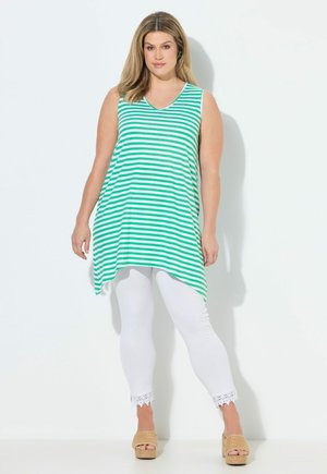 Frau trägt ärmellose grün-weiß gestreifte Tunika mit weißen Spitzenleggings und beigen Plateausandalen, steht vor weißem Hintergrund.