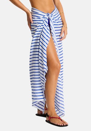 Frau trägt einen blau-weiß gestreiften Sarong, der an der Taille gebunden ist und einen Beinschlitz zeigt, kombiniert mit schwarzen Sandalen mit roten Riemen.