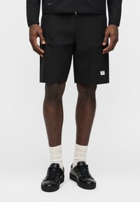 Άνδρας που φοράει μαύρα cargo σορτς με λογότυπο σε ύφασμα στο αριστερό πόδι, λευκές κάλτσες και μαύρα χαμηλά αθλητικά παπούτσια σε απλό φόντο.