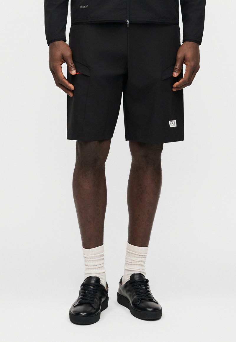 Άνδρας που φοράει μαύρα cargo σορτς με λογότυπο σε ύφασμα στο αριστερό πόδι, λευκές κάλτσες και μαύρα χαμηλά αθλητικά παπούτσια σε απλό φόντο.