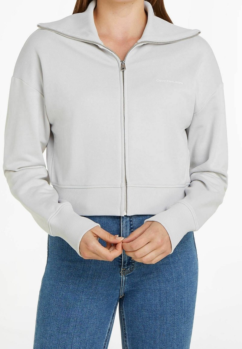 Ljust grå kort sweatshirt med hög krage, dragkedja fram och ribbad nederkant och ärmar. Har subtil logotypdetalj på bröstet.