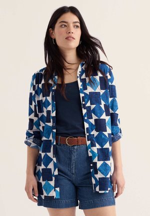 Jonge vrouw met lang donker haar, gekleed in een blauw shirt met geometrisch patroon, donkerblauwe tanktop, spijkershorts en een bruine riem, staat voor een effen achtergrond.