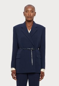 Ναυτικό oversize σακάκι με κοφτές πέτες, μπροστινές τσέπες, μέση με ζώνη και λεία υφή. Από κάτω, εμφανίζεται ένα ελα φρύ πουκάμισο στον γιακά.