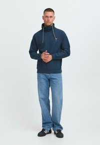 Uomo in piedi con le mani incrociate, indossa un maglione blu scuro a collo alto, jeans azzurri e sneakers nere Adidas con strisce bianche.