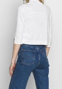 Pullover bianco corto con maniche a 3/4 e dettagli in bottone, abbinato a jeans in denim blu a vita alta con un design di tasche classico.