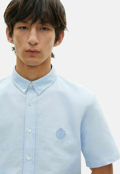 Camisa de botones de manga corta color azul claro con cuello, que presenta un emblema bordado en el lado izquierdo del pecho. Textura de tela suave.