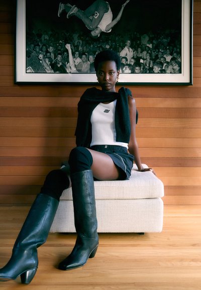 Bottes en cuir noire hauteur genou, débardeur blanc à rayures avec logo, veste noire enroulée autour des épaules, et une jupe noire avec des bordures blanches, assise sur un ottoman de couleur claire.